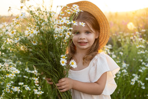 Hermosa niña sonriente con sombrero de paja sostiene ramo de manzanilla de campo al atardecer