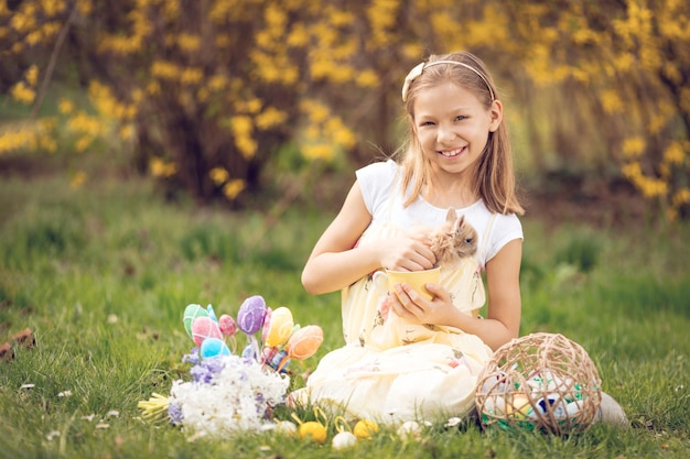 Hermosa niña sonriente sosteniendo un lindo conejito y sentada en el césped con huevos de Pascua y flores en las vacaciones de primavera. Mirando a la cámara.