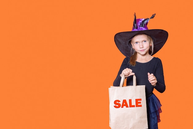 Una hermosa niña sonriente con un vestido negro y un sombrero de bruja sostiene una bolsa de papel artesanal con las palabras VENTA sobre un fondo naranja. Concepto de compras de Halloween