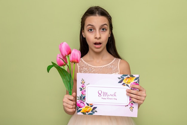 Hermosa niña sorprendida en el día de la mujer feliz sosteniendo flores con tarjeta de felicitación