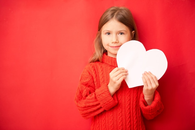 Hermosa niña sosteniendo gran corazón de papel, sobre fondo rojo.