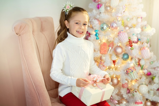 Hermosa niña con un suéter blanco se sienta cerca del árbol de Navidad con un regalo en la mano