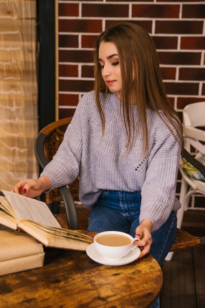 Una hermosa niña con un suéter de punto gris bebe café y lee un libro en una acogedora cafetería, el concepto de ocio y comunicación placenteros. Estudiante o empresaria.