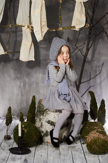 Una hermosa niña de tres años en un traje gris cerca de un cáñamo con decoraciones antiguas.