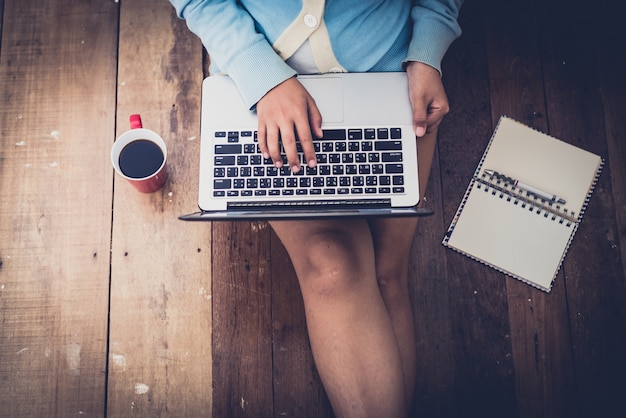 Hermosa niña usa laptop y taza de café en manos de la niña sentada en un piso de madera