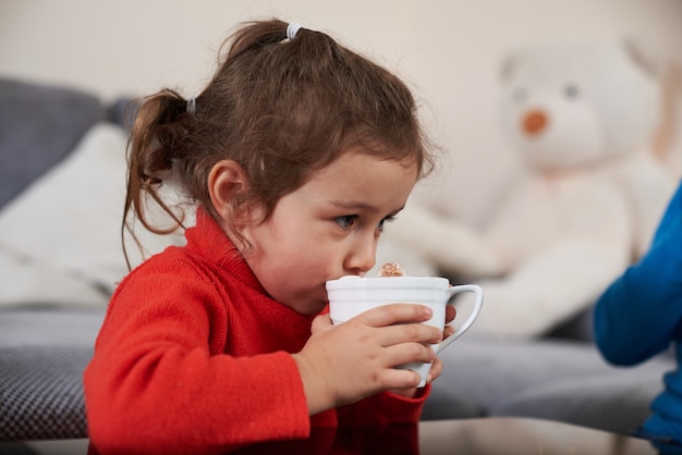 Foto una hermosa niña vestida con un suéter rojo bebiendo un delicioso chocolate con malvaviscos.