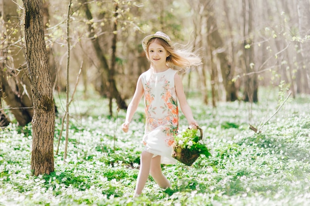 Hermosa niña con un vestido blanco corre en el bosque. Retrato de una niña bonita con un sombrero en la cabeza y una canasta con campanillas.