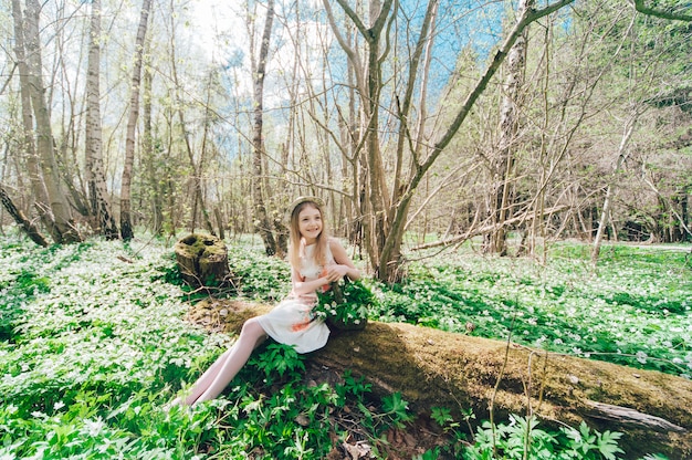 Hermosa niña en un vestido blanco sentado en un árbol caído. Retrato de una niña bonita con un sombrero en la cabeza y campanillas.