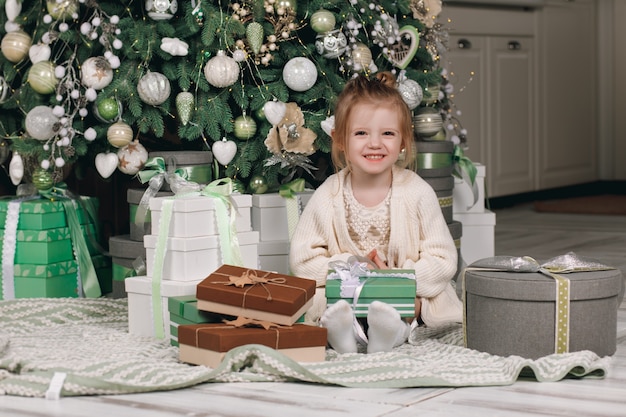 Hermosa niña en un vestido con un regalo en la mano sentada cerca del árbol de Navidad