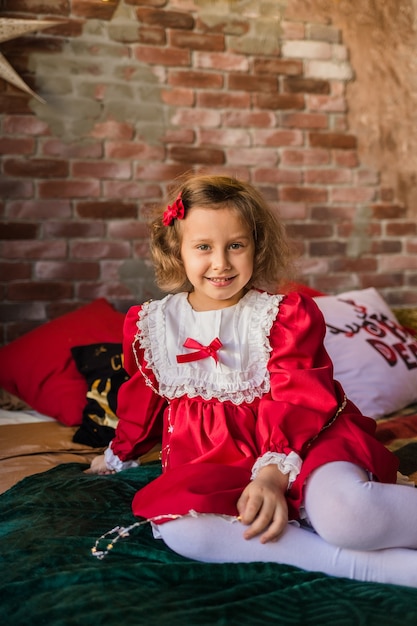 Una hermosa niña con un vestido rojo de fiesta se sienta en la cama en el interior del año nuevo