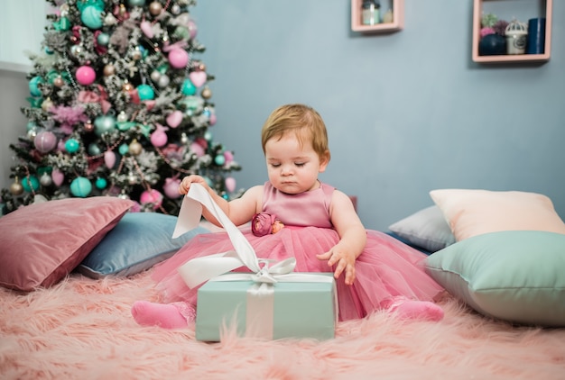 Una hermosa niña con un vestido rosa festivo se sienta sobre una manta de piel y abre un regalo azul en el fondo de un árbol de Navidad