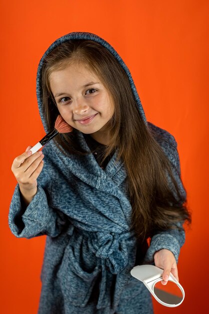 Hermosa niñita de cabello oscuro en bata se para en una bata con un espejo en sus manos aislada en un fondo liso piel de bebé limpia