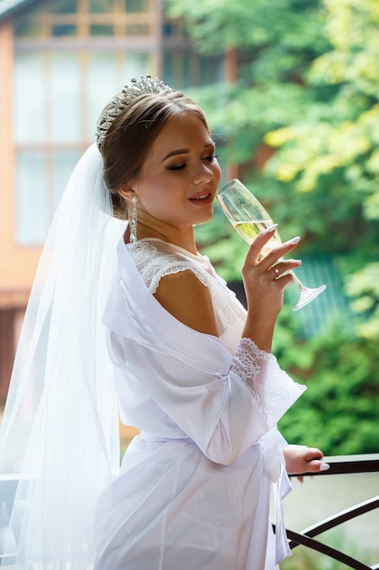 Hermosa novia con una bata blanca en el balcón bebe champán de una copa