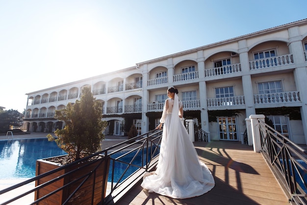 Hermosa novia con cabello oscuro en un lujoso vestido de novia posando en un balcón con vista al mar