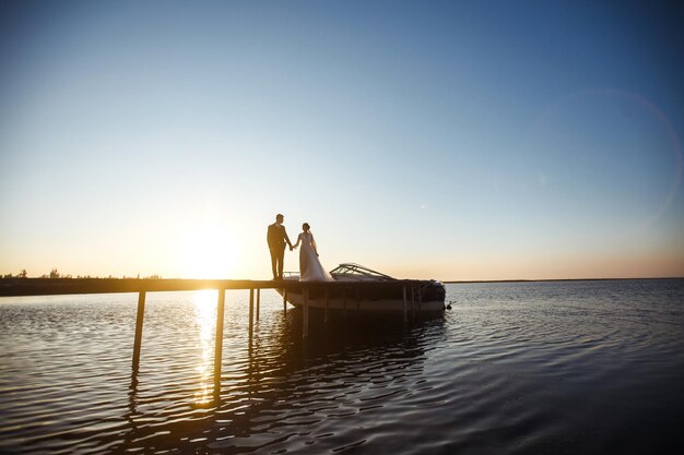 Hermosa novia y elegante novio juntos en el puente contra el fondo del barco al atardecer