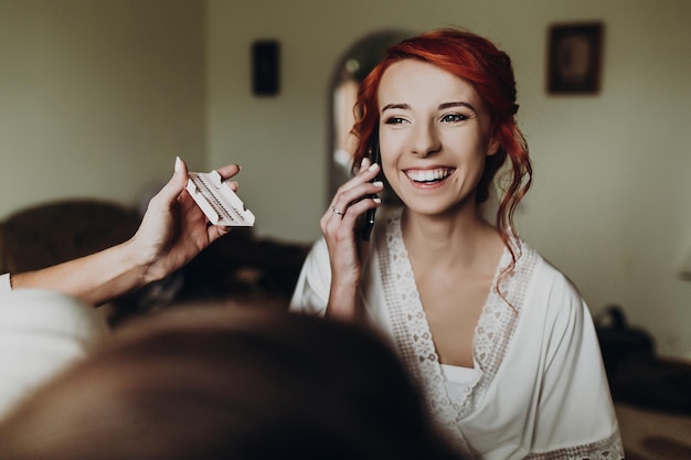 Foto hermosa novia elegante con el pelo rojo hablando por teléfono y sonriendo vistiendo una túnica maquillada por una preparación profesional matutina boda preparando espacio para el texto