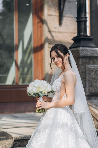 hermosa novia se encuentra en un maravilloso vestido con un ramo en sus manos cerca de una gran casa gris