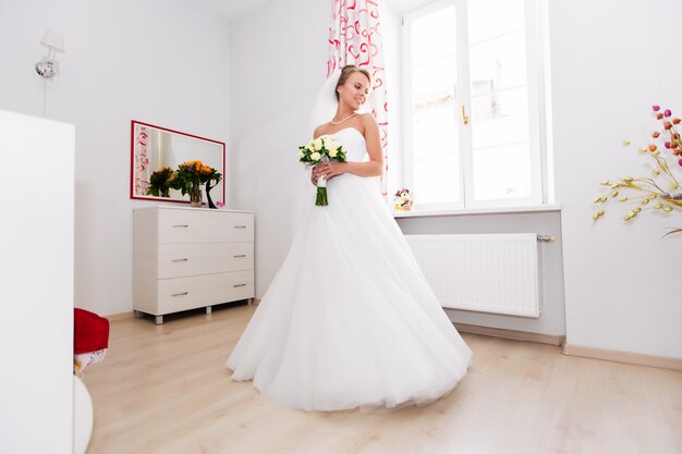 Hermosa novia está de pie junto a la ventana, el día de la boda