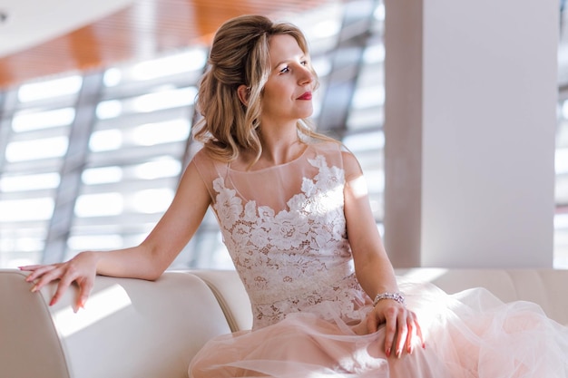 Hermosa novia joven con estilo en un vestido de novia y con un peinado para cabello largo y rubio posando sentada en una silla Los rayos del sol caen sobre la chica desde la ventana