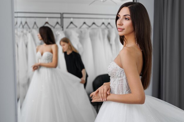 Una hermosa novia joven que se está probando un vestido de novia blanco en una tienda se refleja en el espejo.