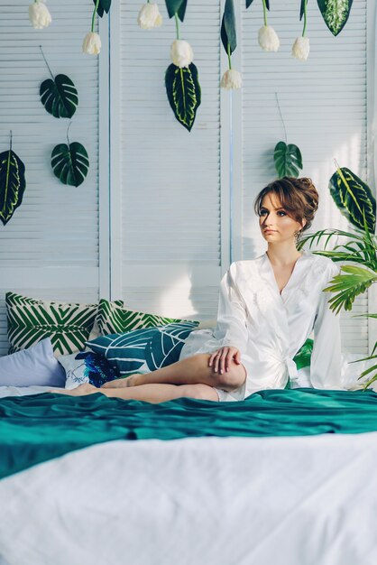 Hermosa novia joven sentada en una cama cubierta con una manta verde en una habitación con flores