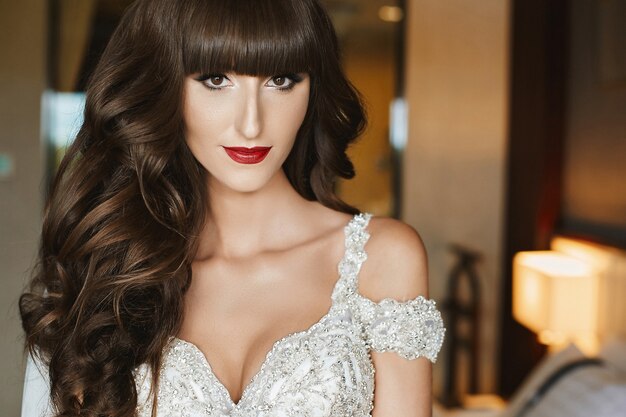 Hermosa novia con maquillaje de novia y cabello largo ondulado en vestido de novia. Modelo de moda en elegante vestido de novia posando en el interior. Mujer joven en vestido de lujo decorado con cristales. Moda de la boda