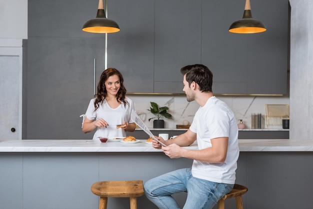 Hermosa novia mirando a un hombre guapo sosteniendo un periódico en la cocina