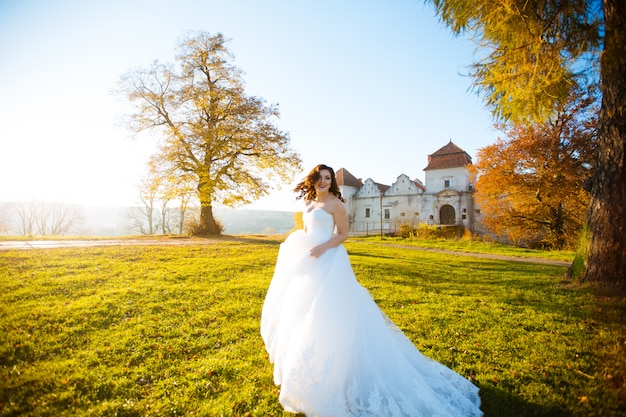 Hermosa novia morena iluminada por el sol en vestido blanco posando en el campo al atardecer con árboles en el fondo