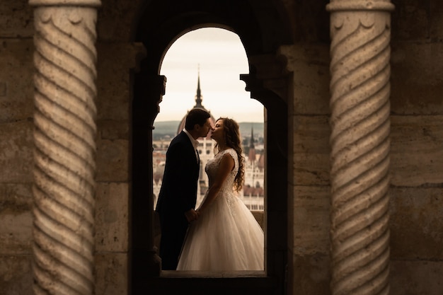 Hermosa novia morena recién casada en vestido de novia blanco y el novio en traje negro posando cerca del antiguo edificio de la columna en Budapest