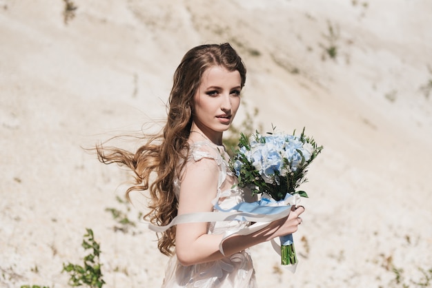 Hermosa novia morena con rizos volando en la montaña de arena en un elegante vestido y un ramo azul y blanco. Boda hermosa