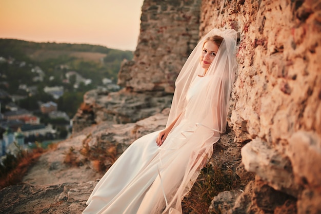 Hermosa novia morena con un vestido de novia y un velo largo posando cerca de las montañas en las afueras de la ciudad. Novia hermosa romántica en un vestido de lujo posando cerca de las rocas.