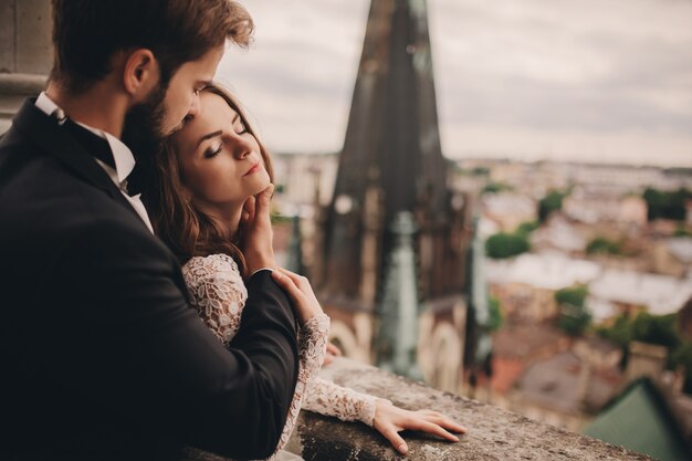 Hermosa novia y el novio elegante se abrazan en el balcón de la antigua catedral gótica con vistas panorámicas de la ciudad