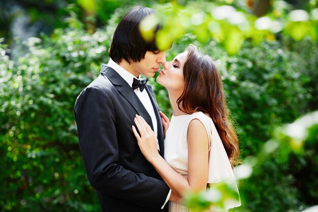 Hermosa novia con el pelo largo y rizado y el novio de pie cerca uno del otro en el fondo de hojas verdes, foto de boda, mirando a los ojos.