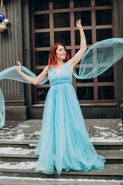 Foto hermosa novia con el pelo rojo con un vestido largo azul en el fondo de la ciudad