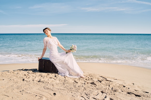 Hermosa novia en la playa en vestido de novia sentada sobre un tronco en la arena