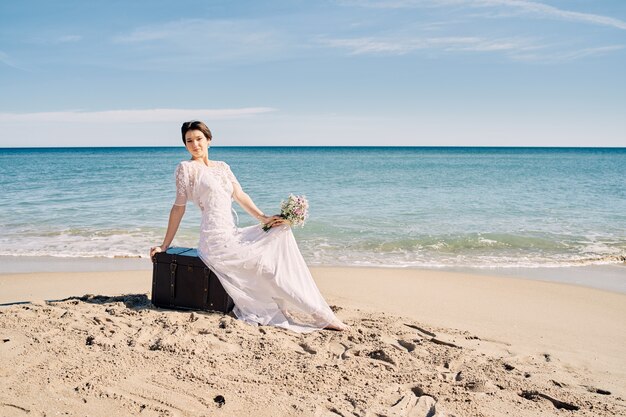 Hermosa novia en la playa en vestido de novia sentada sobre un tronco en la arena