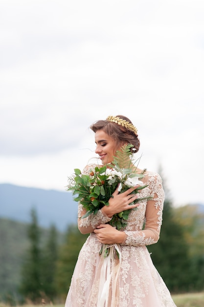 Hermosa novia posando en su vestido de novia en un fondo de montañas. En sus manos sostiene un ramo de flores silvestres.