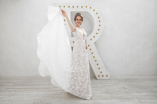Hermosa novia posando en vestido de novia en un estudio fotográfico blanco. Espejo. Sofá. Ramo. La puerta. Candelabro.