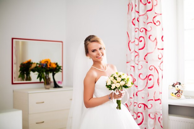 Hermosa novia rubia con vestido de lujo blanco se está preparando para la boda. Preparaciones matutinas. Mujer poniéndose el vestido.