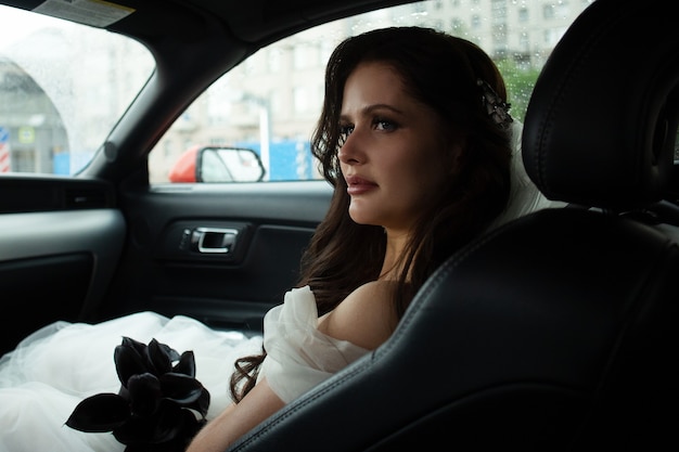 La hermosa novia está sentada en un coche rojo con un vestido blanco y un ramo de flores. concepto de boda