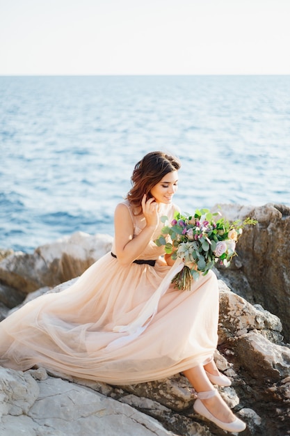 Hermosa novia sonriente con un vestido beige con un ramo de flores en sus manos se sienta sobre una roca arriba