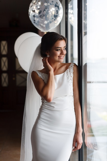 Hermosa novia sonriente en vestido de novia de moda interior cerca de la ventana