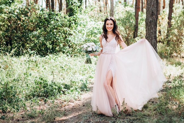 Hermosa novia en un tierno vestido rosa al aire libre, niña sostiene un ramo, una niña con un vestido largo camina en un bosque de pinos