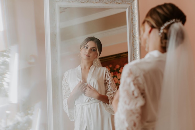 Hermosa novia en túnica posando junto a la ventana, preparación de la boda