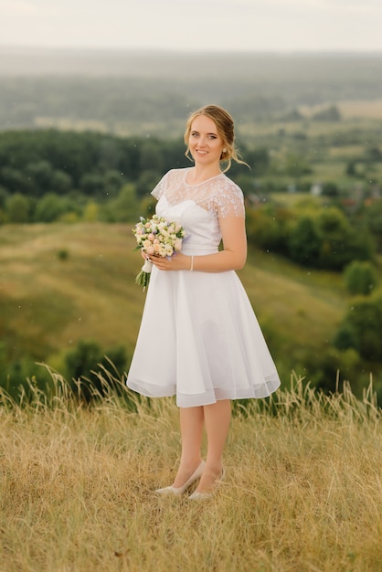 Una hermosa novia con un vestido blanco está en la naturaleza y tiene un ramo de novia en sus manos.