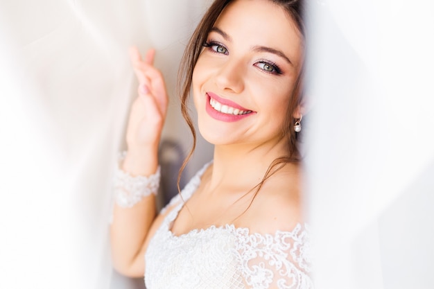 Hermosa novia en vestido blanco mira a cámara y sonríe bajo la cortina