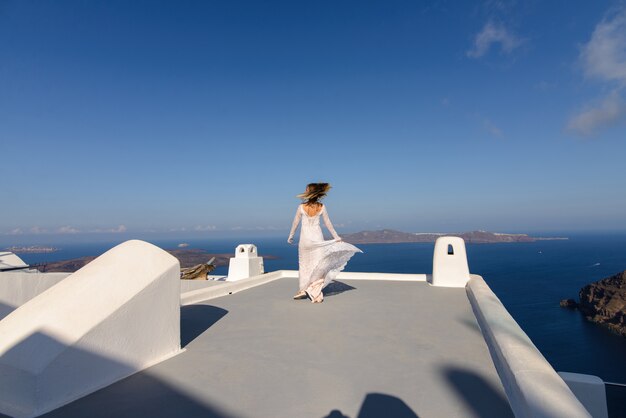 Hermosa novia con un vestido blanco posando en el techo de la casa contra el fondo del mar Mediterráneo en Thira, Santorini.