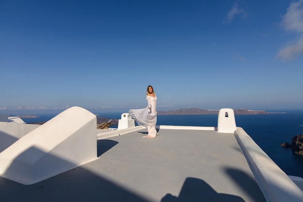 Hermosa novia con un vestido blanco posando en el techo de la casa contra el fondo del mar Mediterráneo en Thira, Santorini.