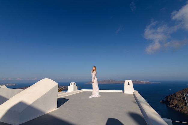 Hermosa novia con un vestido blanco posando en el techo de la casa contra el fondo del mar Mediterráneo en Thira, Santorini.