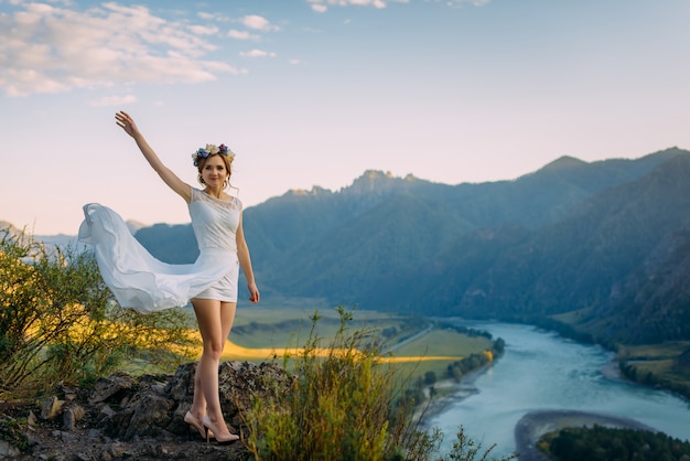 Hermosa novia en vestido corto blanco posando sobre el paisaje de las montañas y el río con azul cielo nublado